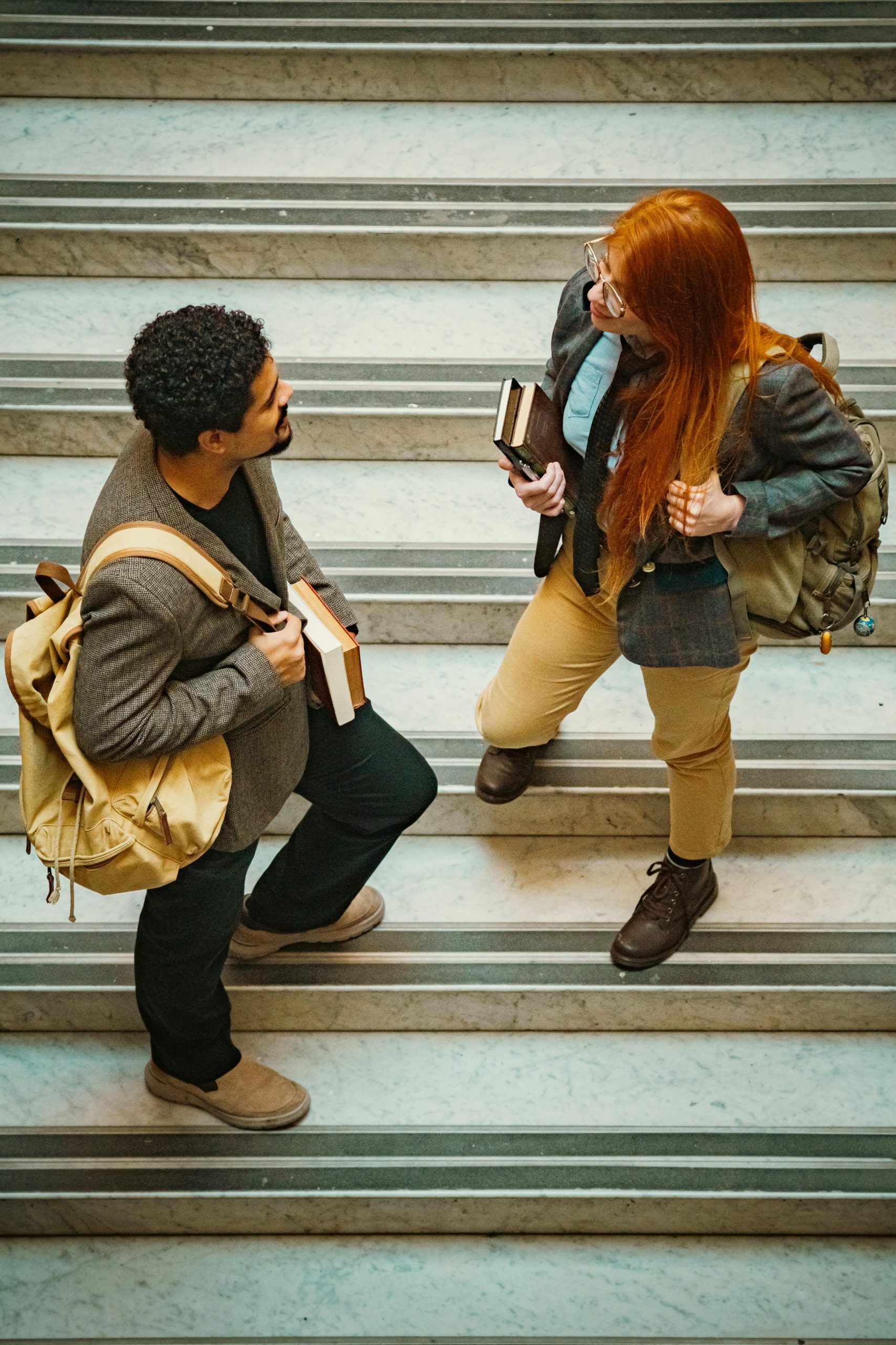 Two college students carrying books and backpacks discuss education financing on a university staircase while researching how to Apply for Student Loan programs, using a Student Loan Interest Calculator, exploring Student Loan Forgiveness for Teachers, comparing Current Student Loan Refinance Rates, and learning how to Refinance Student Loans with a Cosigner.